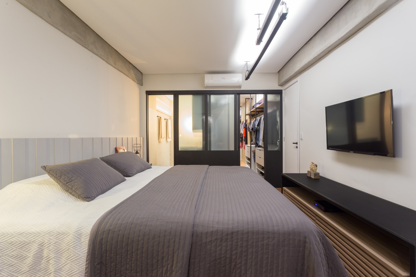 Demolimos as paredes que dividiam o banheiro e o closet do quarto e criamos uma esquadria em
madeira com portas de correr com vidro jateado, que permite melhor iluminar esses espaços além de
contribuir para a linguagem industrial pretendida para o apartamento. A luminária pendente, executada pelos próprios clientes, dá uma iluminação indireta ao ambiente,
criando um clima mais aconchegante.
