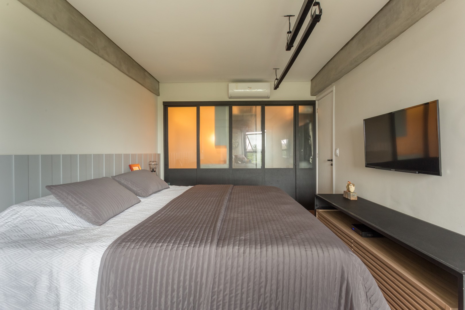 Demolimos as paredes que dividiam o banheiro e o closet do quarto e criamos uma esquadria em
madeira com portas de correr com vidro jateado, que permite melhor iluminar esses espaços além de
contribuir para a linguagem industrial pretendida para o apartamento. A luminária pendente, executada pelos próprios clientes, dá uma iluminação indireta ao ambiente,
criando um clima mais aconchegante.