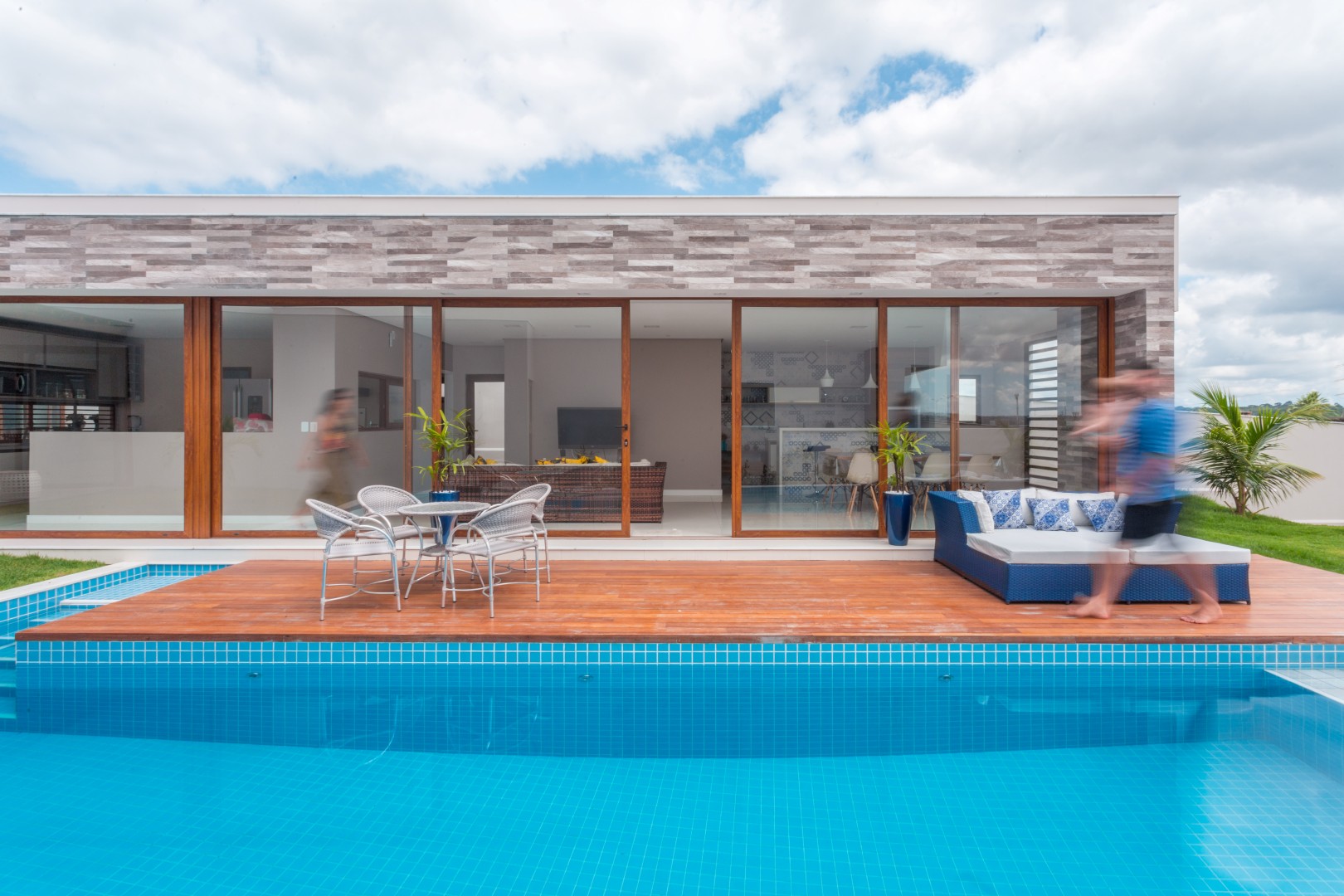 Ao fundo, vê-se a cozinha, que se localiza na área central do volume social, e mantém comunicação visual tanto com a sala de jantar quanto a área de lazer, que por sua vez se integra totalmente ao deck da piscina através das grandes portas de correr em pvc e vidro.