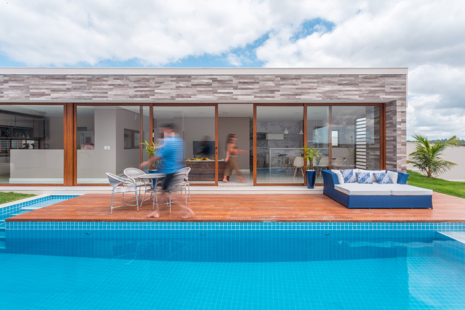 Ao fundo, vê-se a cozinha, que se localiza na área central do volume social, e mantém comunicação visual tanto com a sala de jantar quanto a área de lazer, que por sua vez se integra totalmente ao deck da piscina através das grandes portas de correr em pvc e vidro.