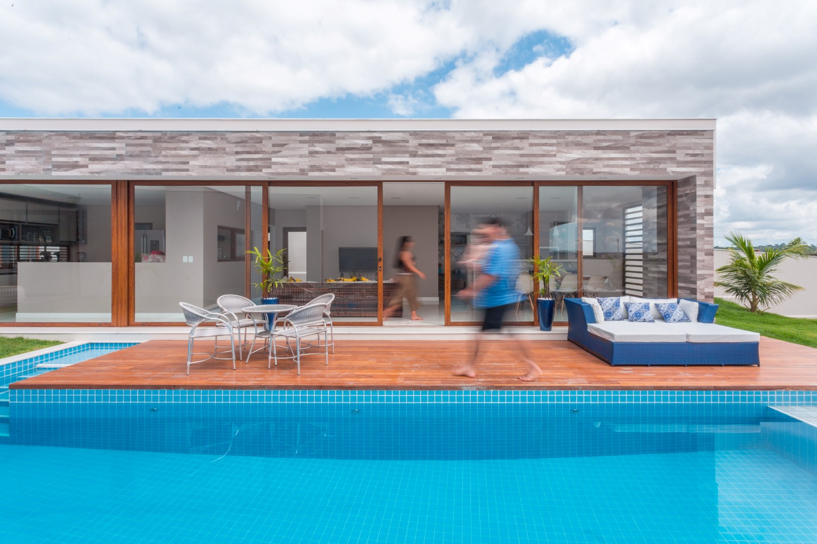 Ao fundo, vê-se a cozinha, que se localiza na área central do volume social, e mantém comunicação visual tanto com a sala de jantar quanto a área de lazer, que por sua vez se integra totalmente ao deck da piscina através das grandes portas de correr em pvc e vidro.
